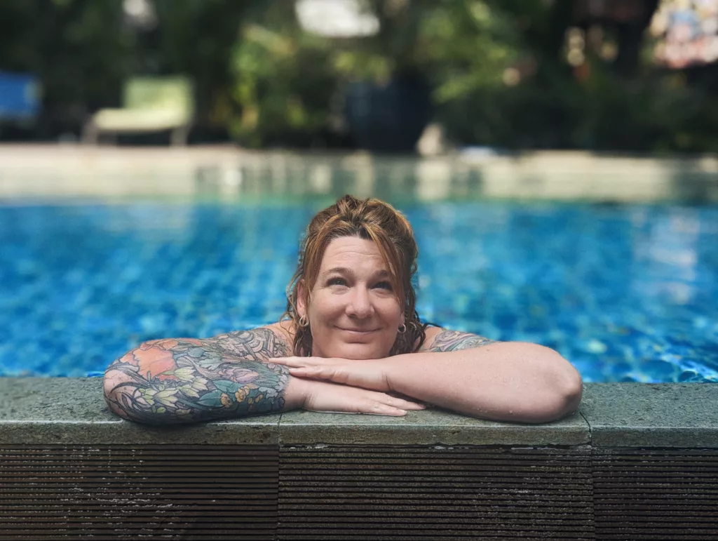 A red-haired woman with colorful sleeve tattoos leans on the edge of a bright blue swimming pool against a blurred tropical background.