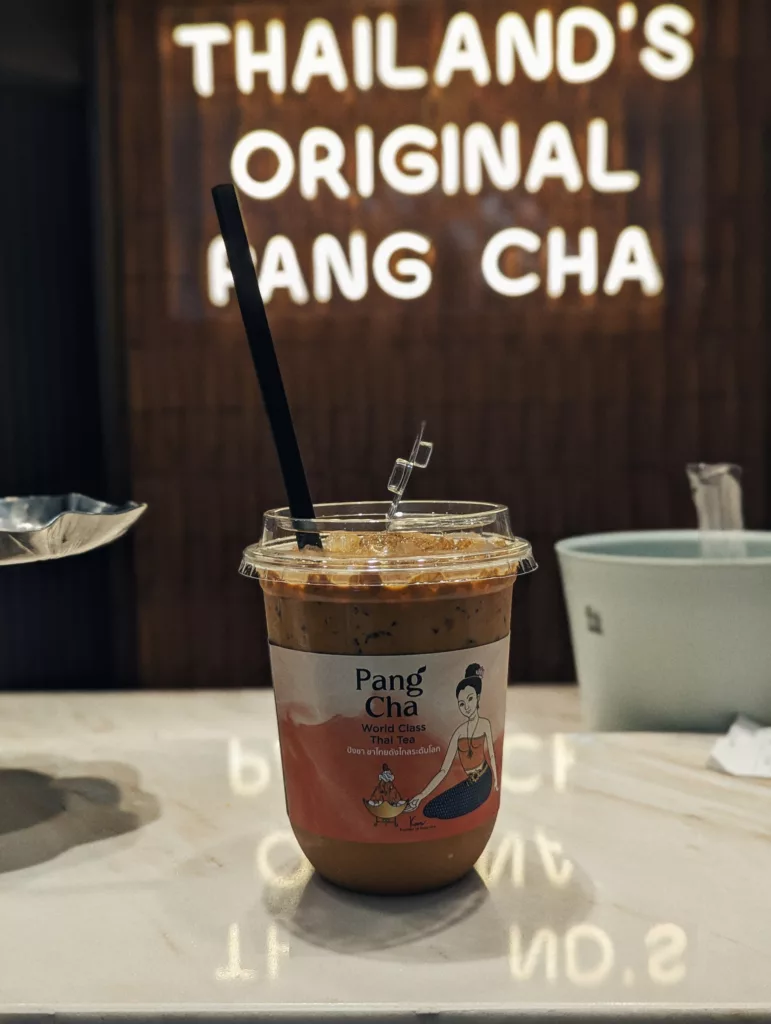 Iced Pang Cha Thai tea with a black straw on a marble counter, set against a glowing neon sign reading THAILAND’S ORIGINAL PANG CHA.