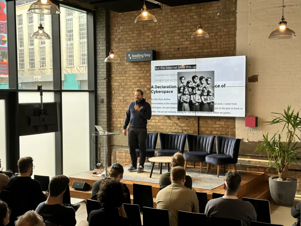 A speaker on a stage in an industrial venue presenting a slide titled "A Declaration of Independence of Cyberspace" to an audience.