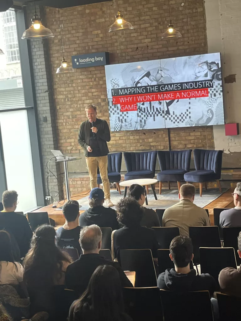 A man presents to an audience at Loading Bay. A screen reads: 1. Mapping the games industry 2. Why I won't make a normal game.