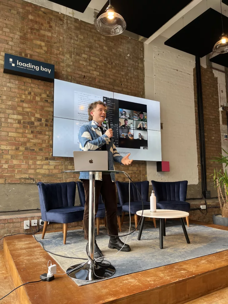 A person presents on a stage at Loading Bay, speaking into a microphone before a large screen showing a video call in a brick-walled room.
