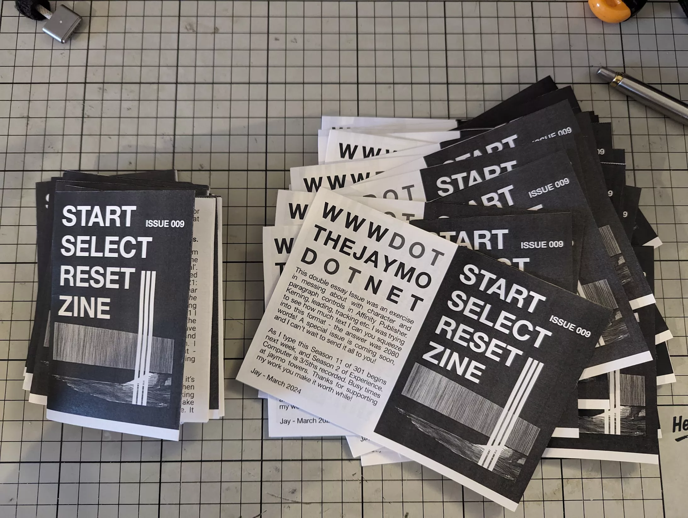 A stack and an open copy of the black and white START SELECT RESET ZINE ISSUE 009 on a grey gridded cutting mat next to a silver pen.