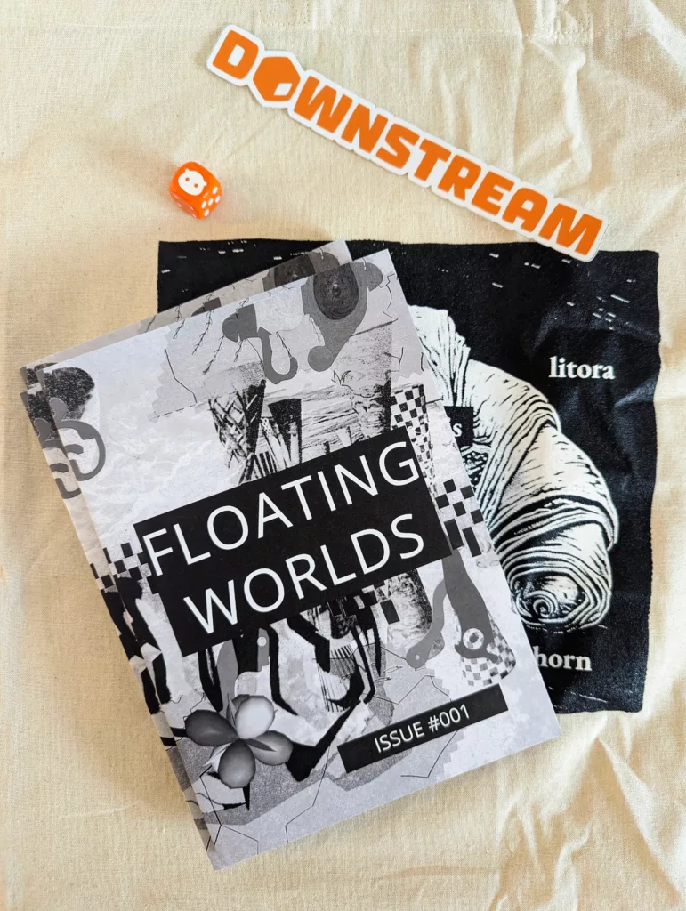 Floating Worlds Issue #001 zine, a black litora horn print, an orange Downstream sticker, and a small orange die on a beige fabric surface.
