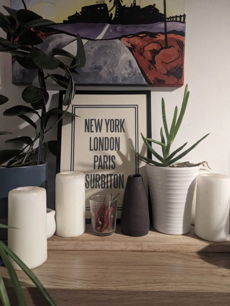 A wooden shelf with plants, white candles, and a framed print reading NEW YORK LONDON PARIS SURBITON beneath a landscape painting.