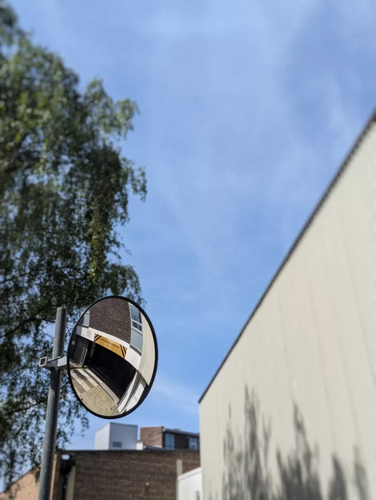 A convex traffic mirror reflects a brick building and car park entrance against a clear blue sky, green tree, and a plain beige wall.