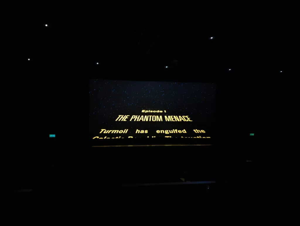 The yellow opening crawl of Star Wars Episode I: The Phantom Menace on a cinema screen in a dark auditorium with green exit signs.