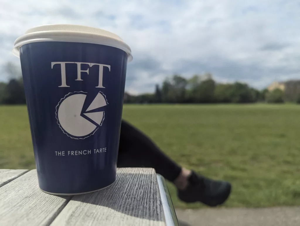 A blue The French Tarte takeaway cup on a wooden table, with a grassy field and blurred person in the background under a cloudy sky.