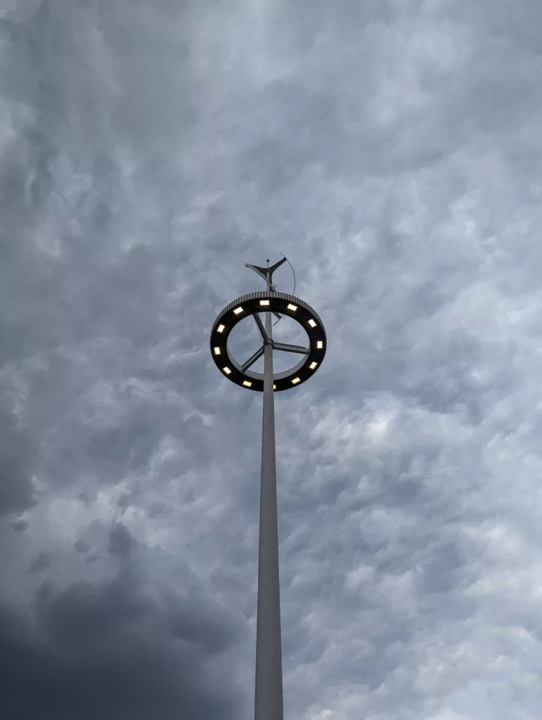 A tall light pole with a glowing circular ring of LEDs topped by a small wind vane against a dark, stormy grey sky.