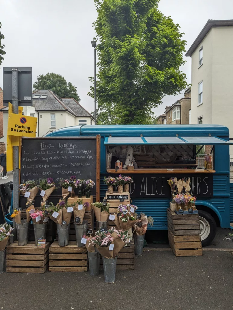 A blue vintage flower van parked on a street with bouquets in paper wraps displayed on wooden crates and a chalkboard listing floral workshops.