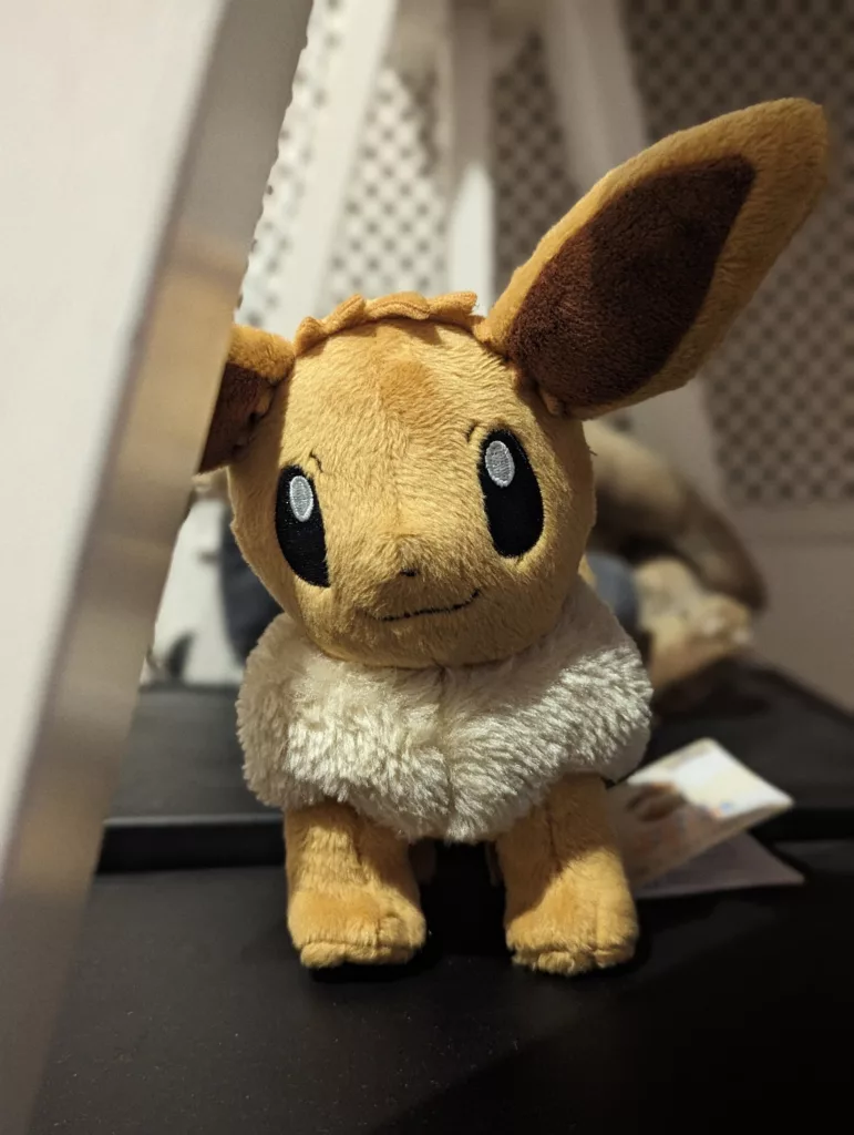 A small plush Pokémon Eevee standing on a black surface, featuring its brown fur, large ears, and a thick white fluffy collar.