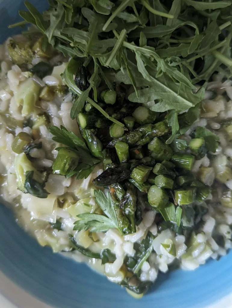 A creamy green risotto topped with charred asparagus tips, fresh parsley, and a handful of arugula in a blue bowl.