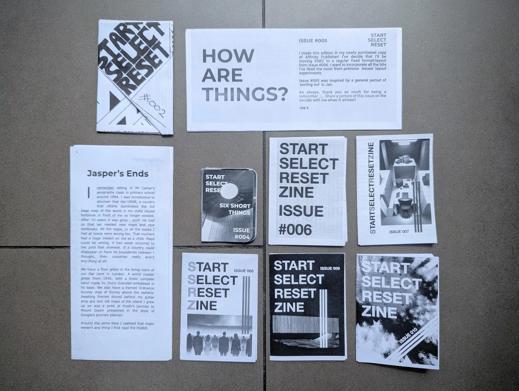 A collection of black and white zines and printed materials from Start Select Reset arranged on a grey tiled floor.