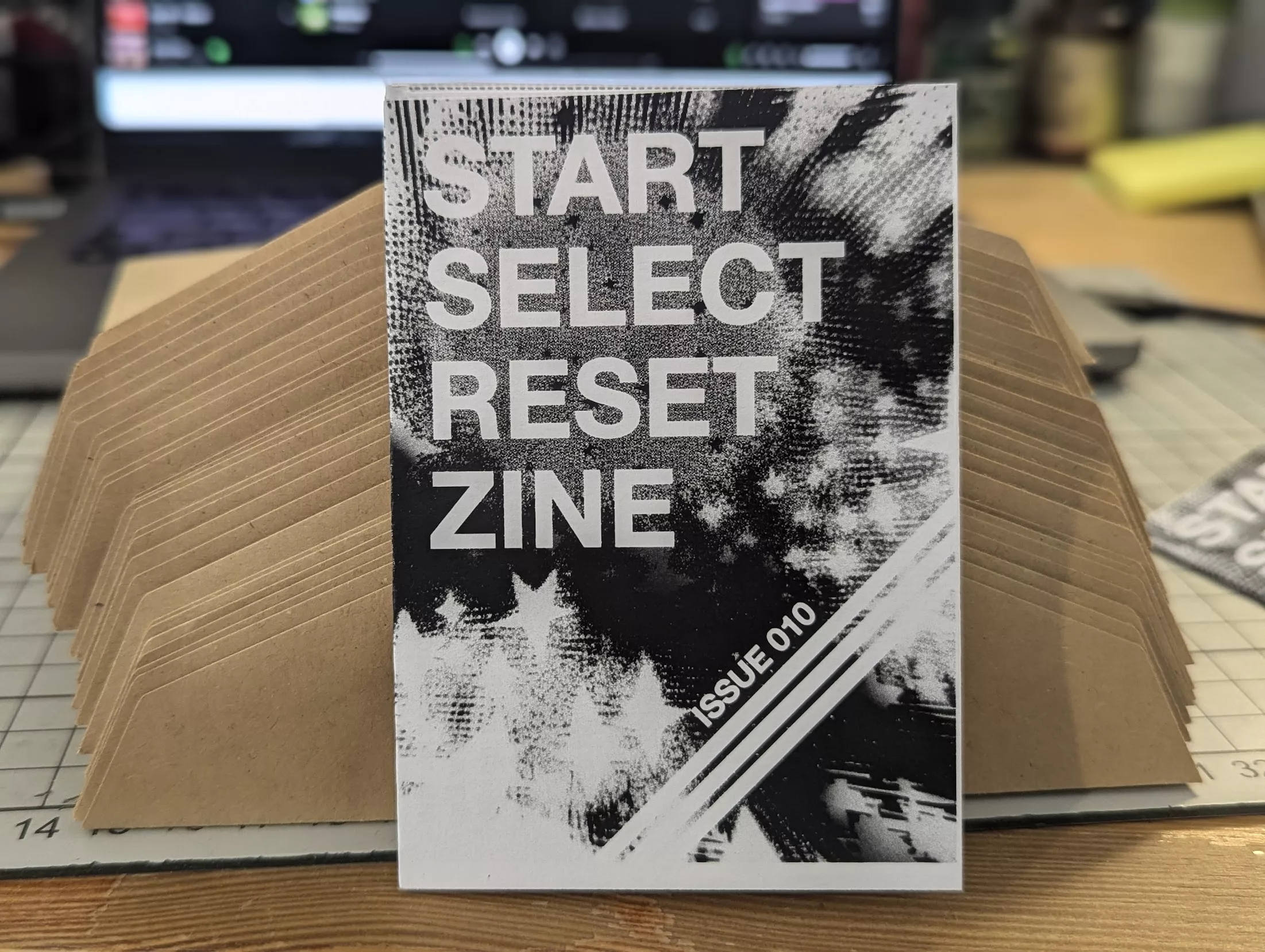 START SELECT RESET ZINE ISSUE 010 with a black and white abstract cover, propped against a fan of brown envelopes on a desk.