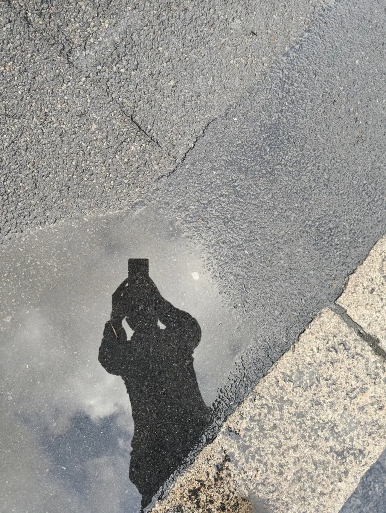 The reflection of a person holding a phone to take a picture in a puddle on a grey asphalt pavement next to a stone curb.