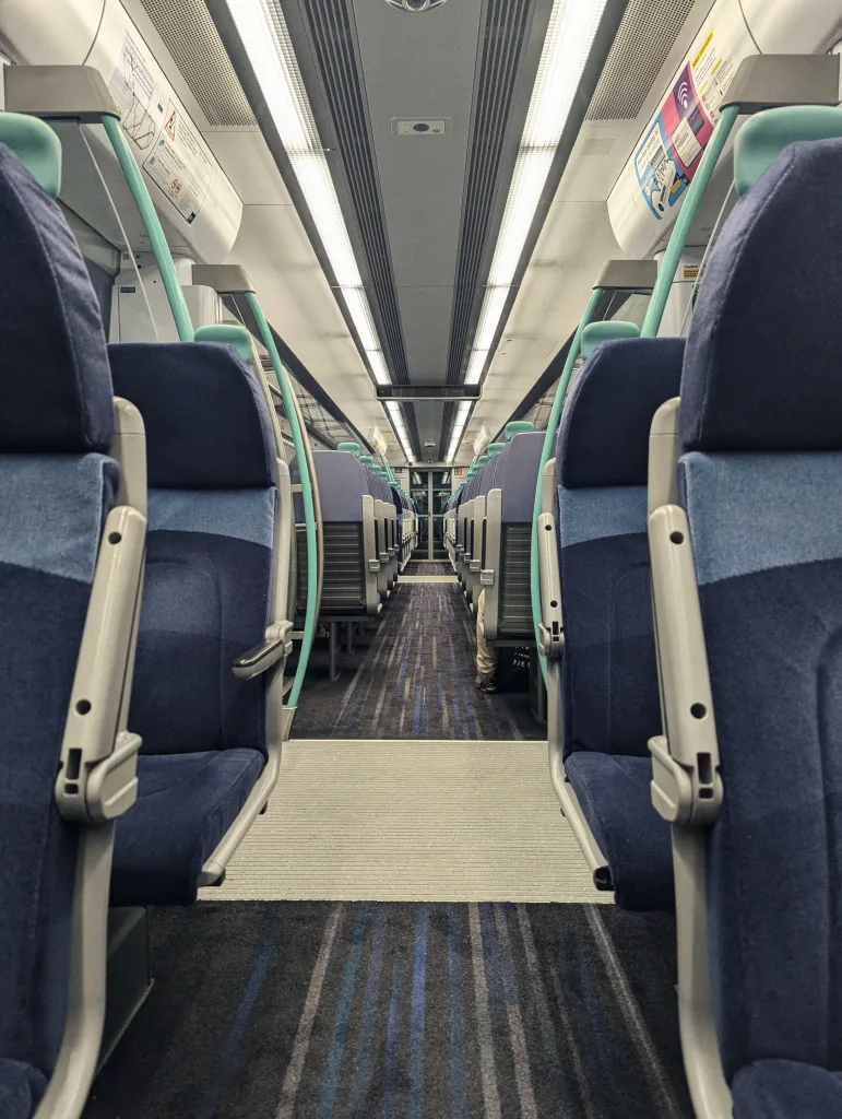 View down the center aisle of a symmetrical train carriage with blue fabric seats, teal handrails, and bright overhead strip lighting.