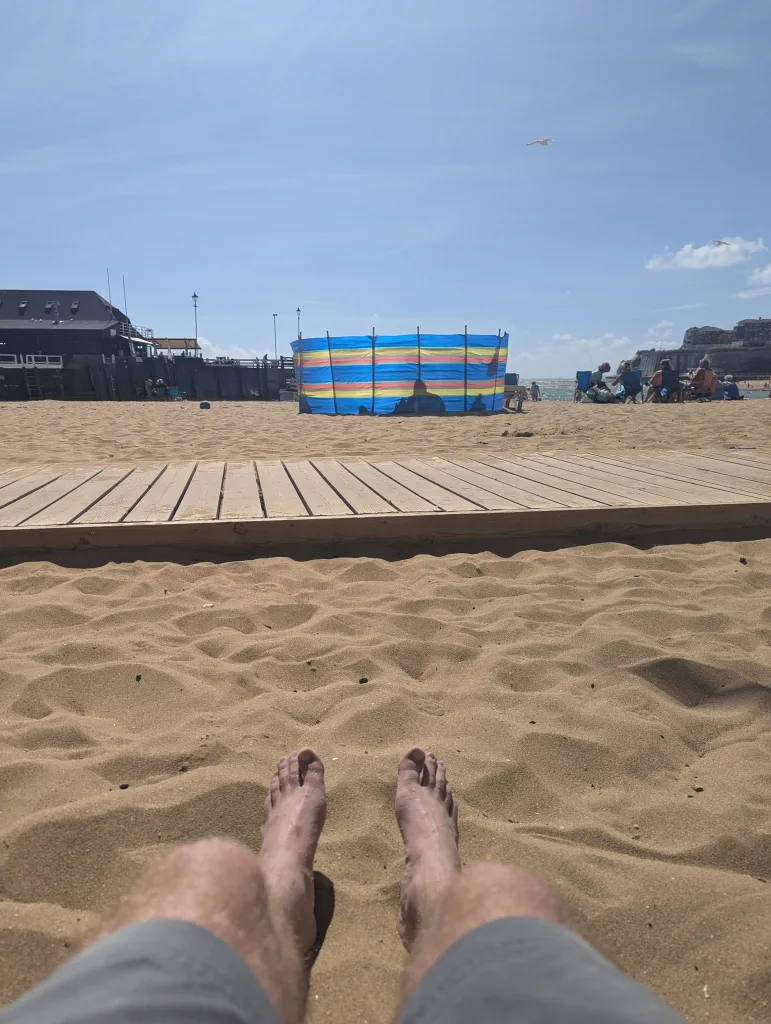 A first-person view of bare feet in the sand, looking towards a wooden walkway and a colourful striped windbreak on a sunny beach.