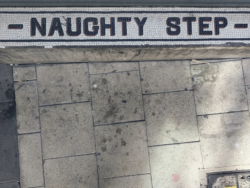 A mosaic tile step reading "NAUGHTY STEP" in black capital letters on a white background above grey paving stones.