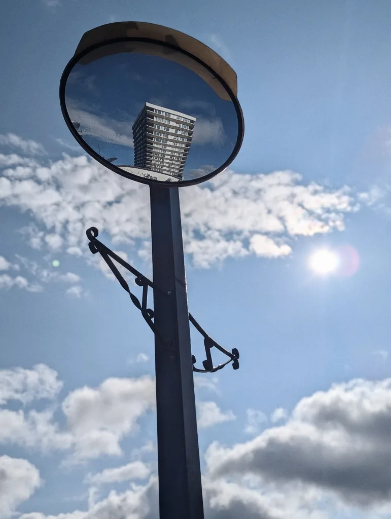 A high-rise tower block reflected in a convex traffic mirror against a bright, cloudy blue sky with the sun shining in the background.