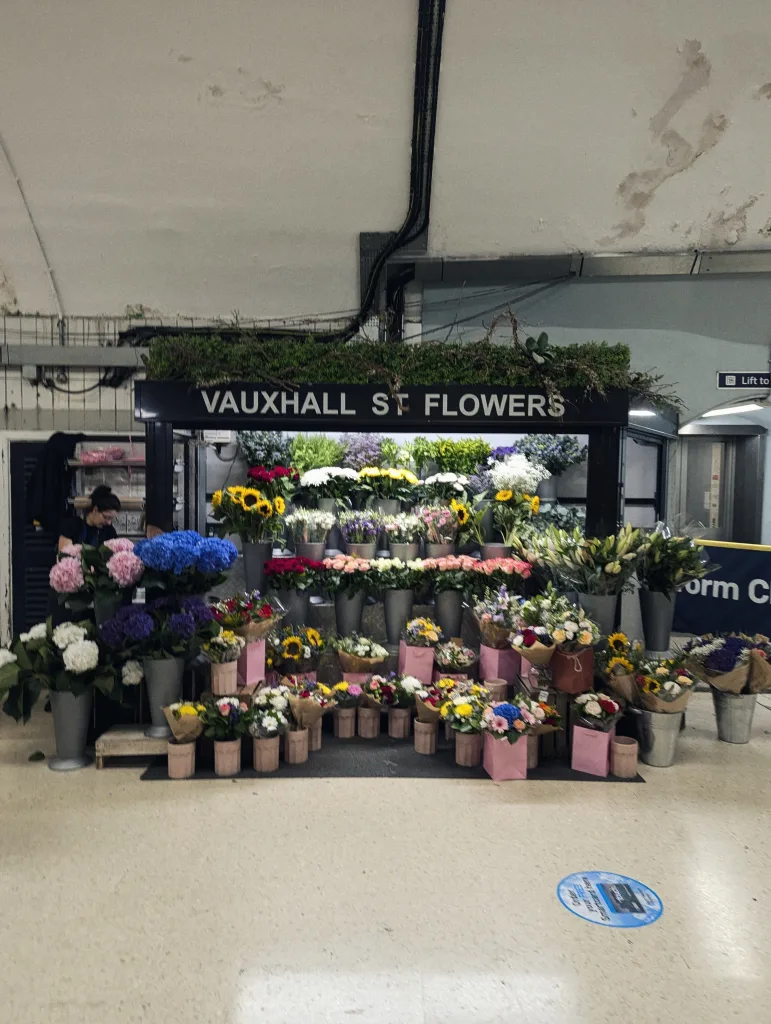 Vauxhall St Flowers stall inside a station, with tiered displays of sunflowers, hydrangeas, and mixed bouquets in buckets and pink bags.