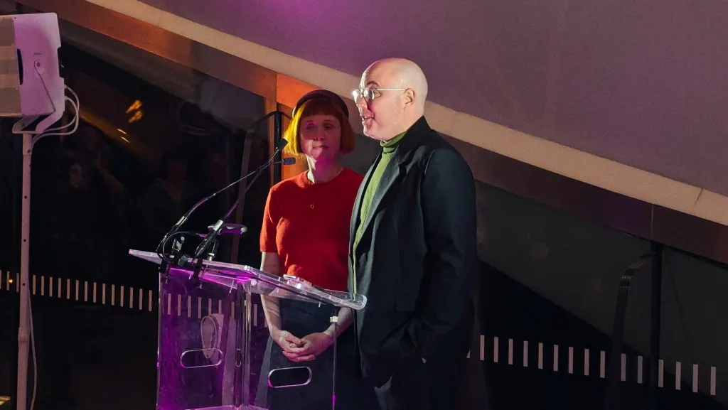 A bald man in a black suit and a woman with orange hair and a red top speaking at a clear acrylic lectern under dim purple lighting.