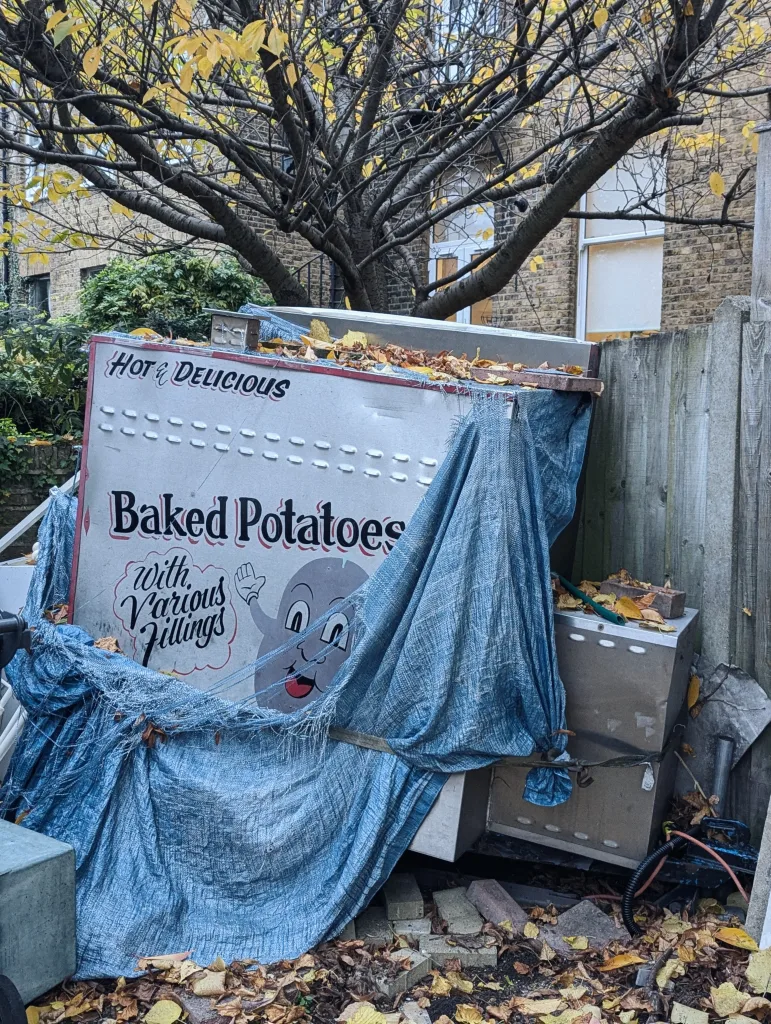 A discarded baked potato food stall partially covered by a blue tarp, nestled under a tree with yellow autumn leaves.