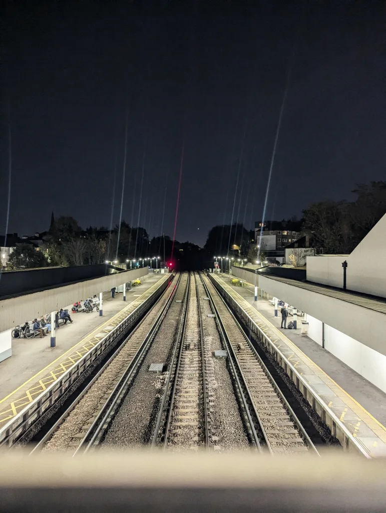 Railway tracks at night between two lit platforms. Bright streaks of light rise into the dark sky from the platform lamps.