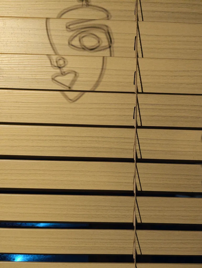 An abstract line drawing of a face with a heart shape and large eye projected onto the slats of light wooden window blinds.