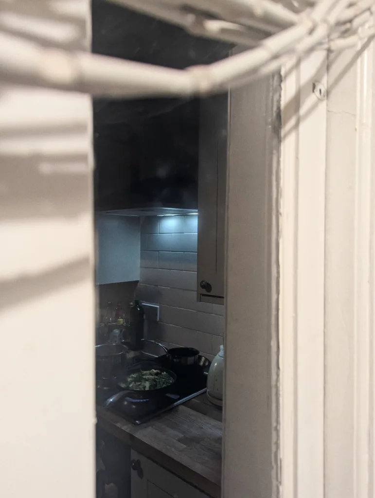 Green vegetables cooking in a pan on a gas stove, seen through a narrow opening between white walls.