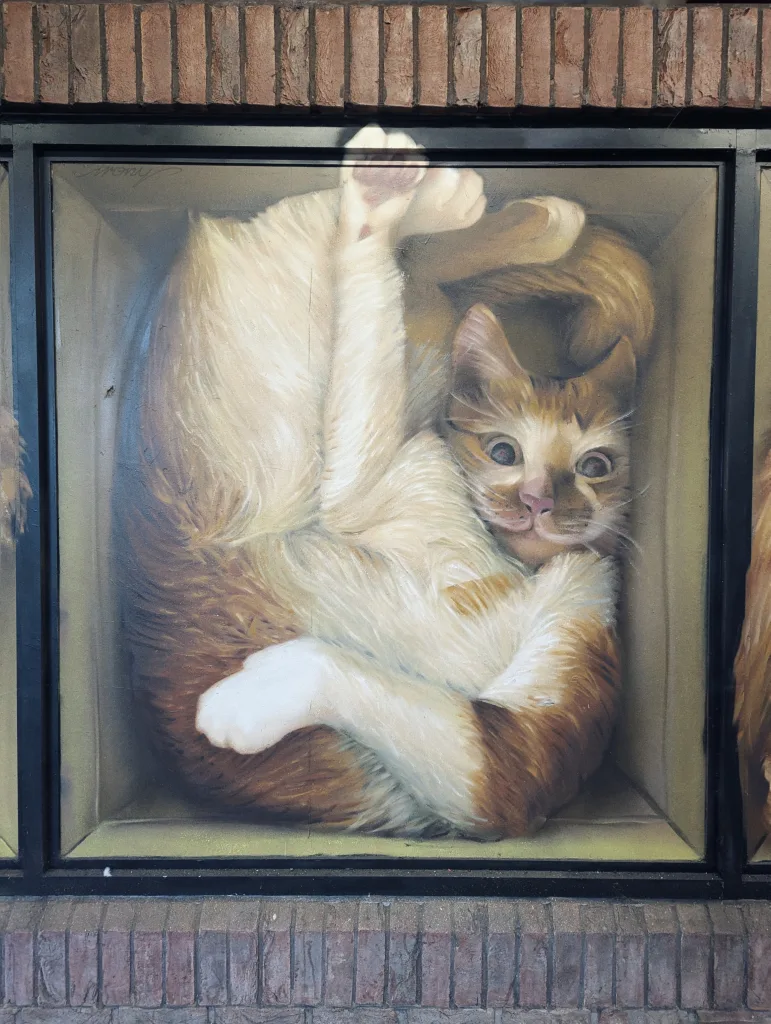 A realistic mural of a ginger and white cat squeezed into a cardboard box, framed by black borders and brickwork.