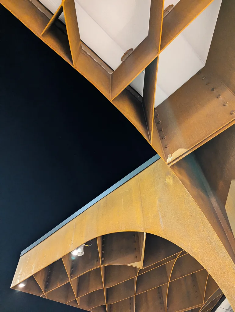 Weathered orange steel girders forming a geometric lattice and archway roof of Kingston bus station against a dark night sky.