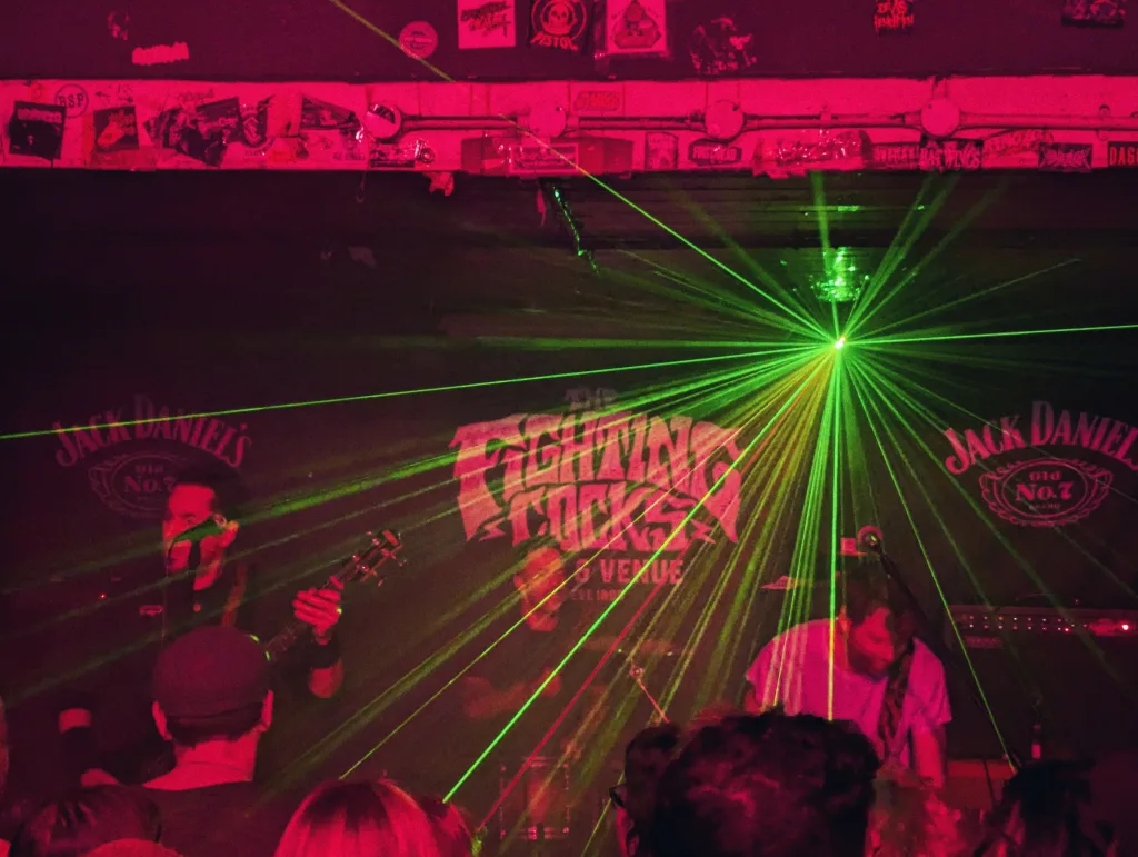Green lasers radiate across a band performing on a red-lit stage at The Fighting Cocks venue, with Jack Daniel's logos on the back wall.