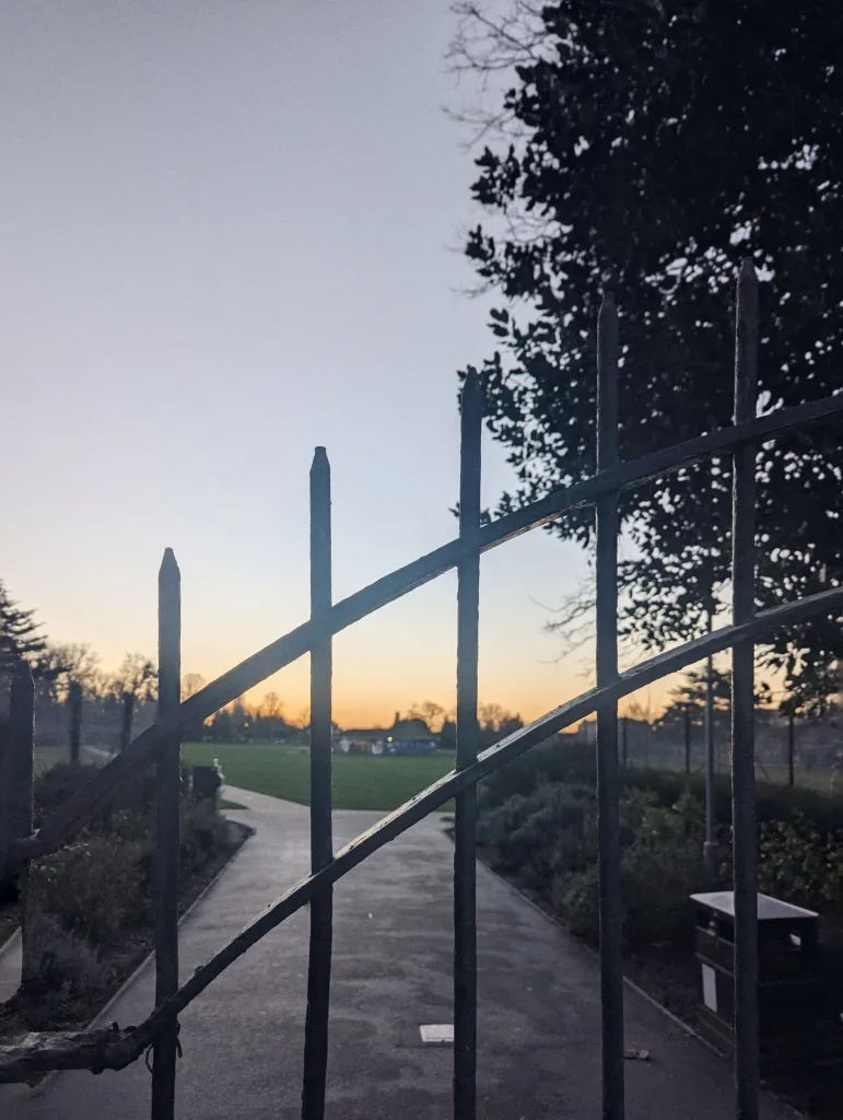 Spiked iron fence in front of a park path leading to a field under a sunset sky with an orange horizon and silhouetted trees.