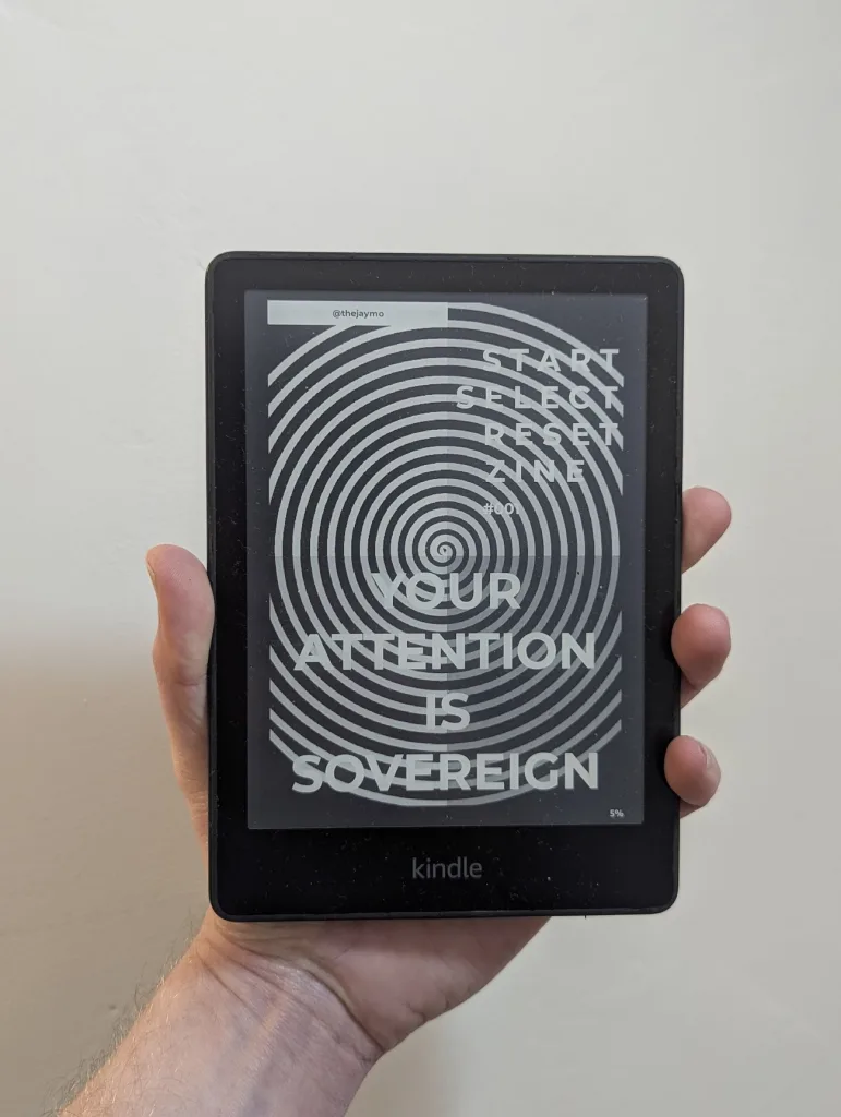 Start Select Reset Zine #001 - A hand holds a Kindle displaying a spiral cover with the text Your Attention is Sovereign.