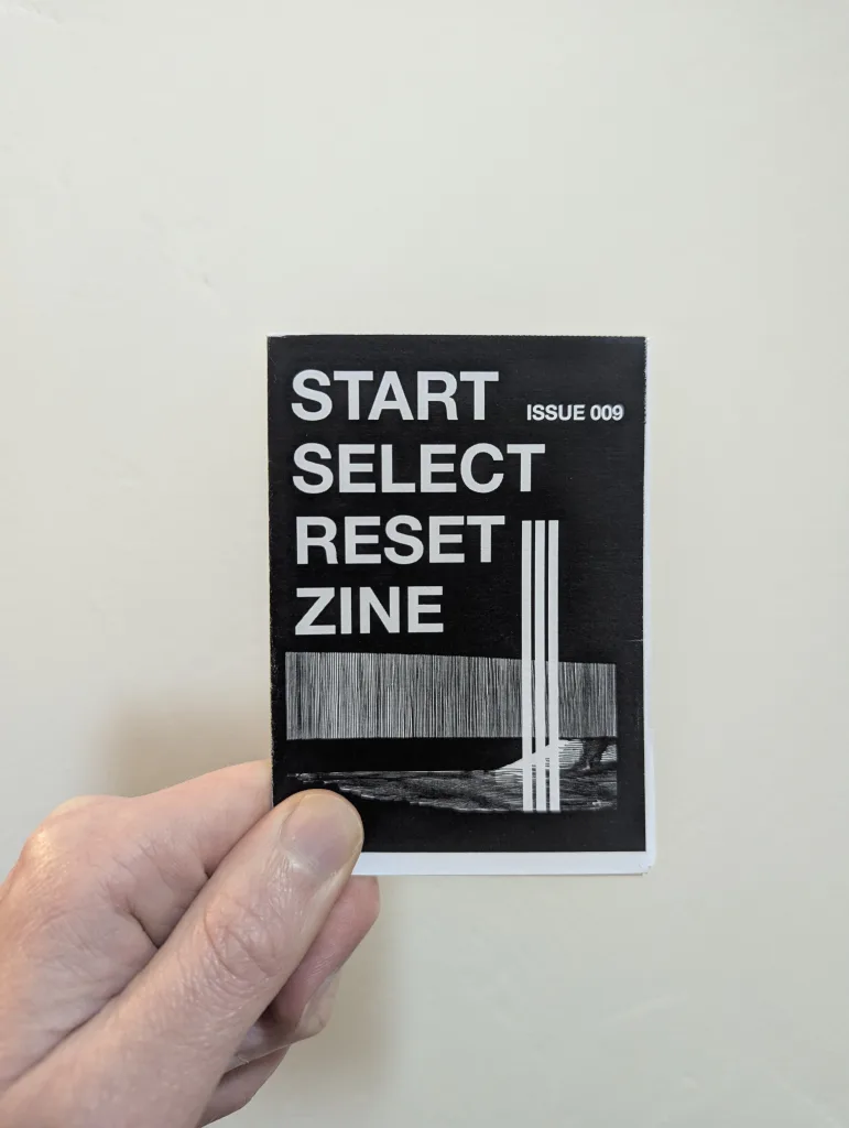 Featured image for Start Select Reset Zine Issue 009 - a hand holding the black and white zine against a plain wall.