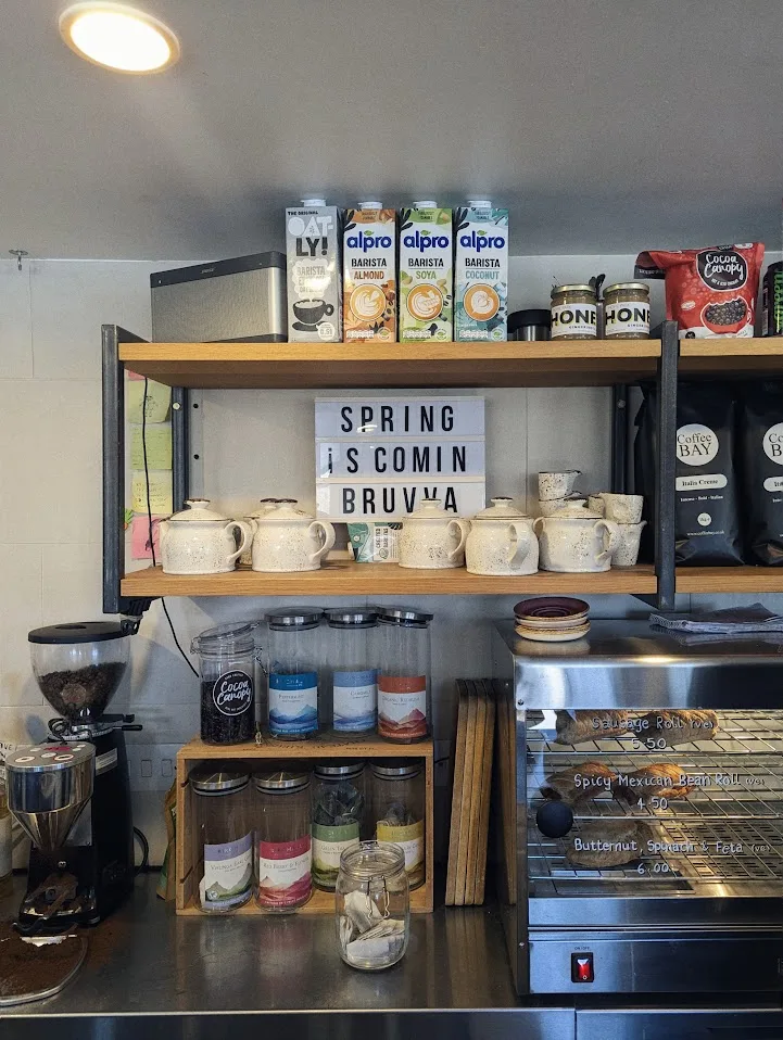 Shelves in a cafe with plant milks, teapots, and a lightbox reading SPRING IS COMIN BRUVVA above a pastry display case.