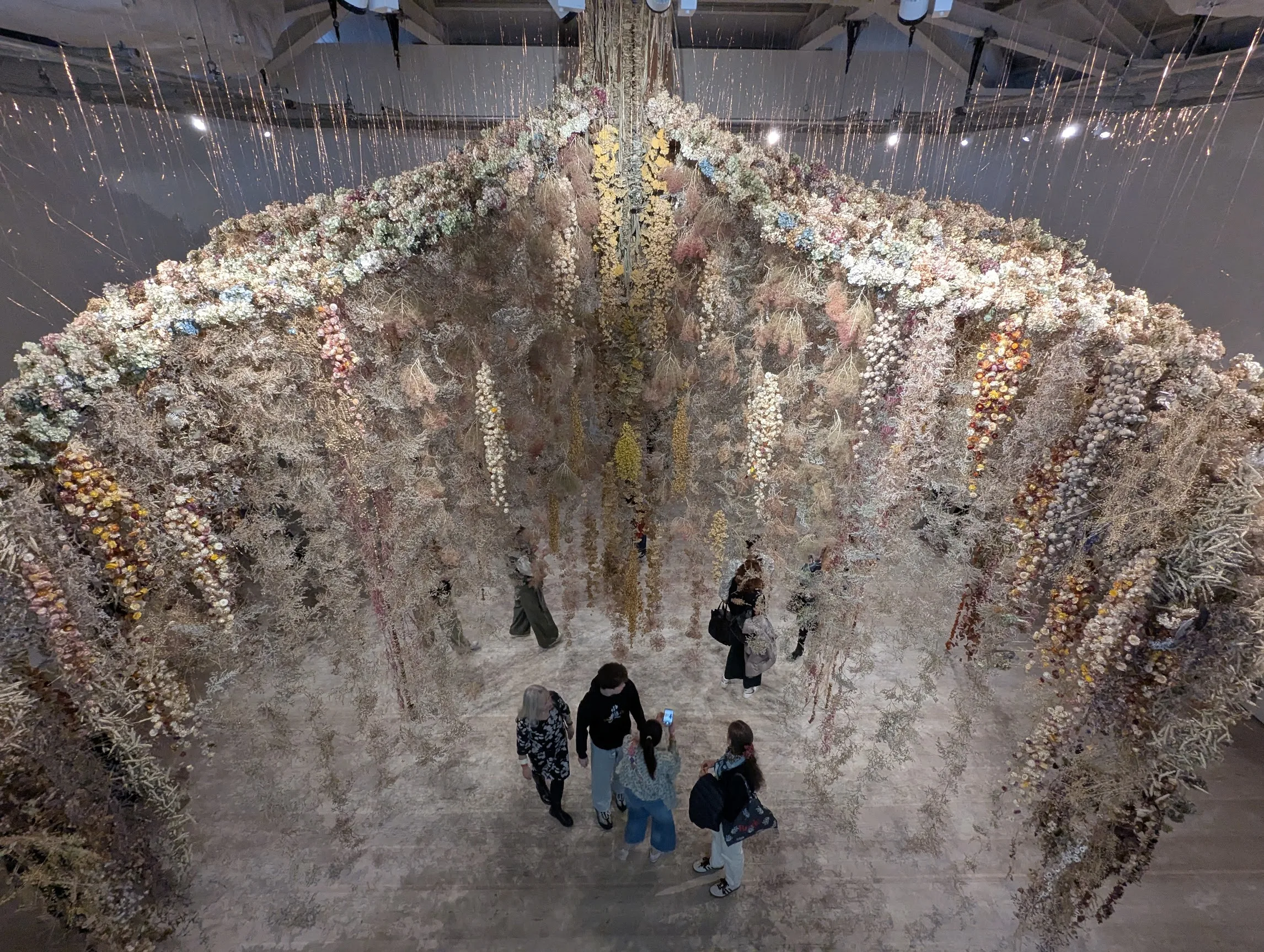 A large-scale installation of suspended dried flowers forming a floral canopy over visitors in a cavernous gallery space.