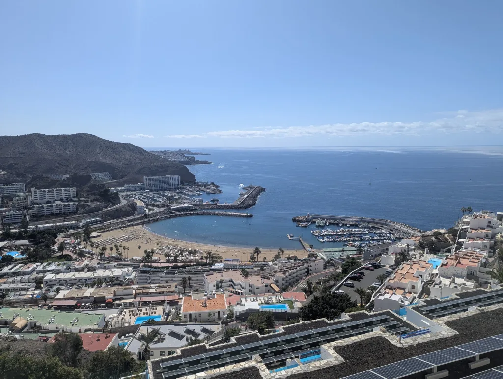 High-angle view of a sunny coastal resort featuring a curved sandy beach, boat-filled marina, and white buildings under a clear blue sky.