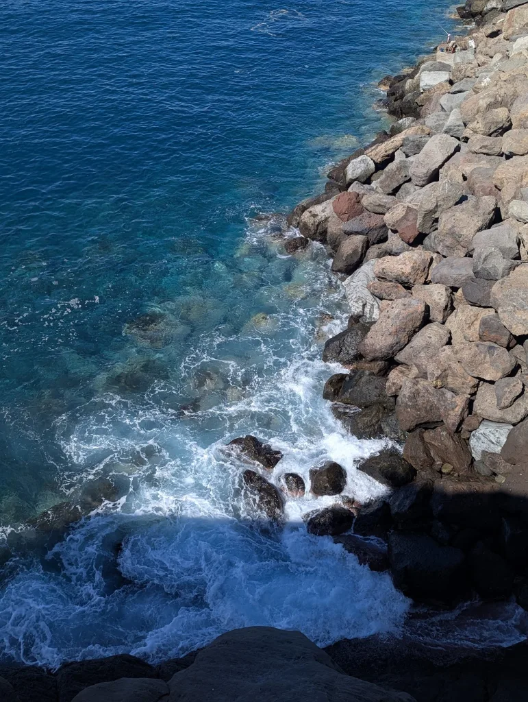 White foam from bright blue ocean waves crashing against a steep bank of large brown and grey boulders along a coastline.