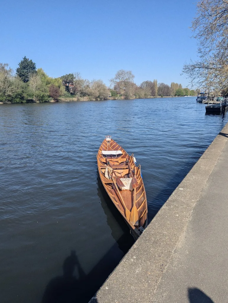 A traditional wooden rowing boat with oars moored beside a concrete path on a wide river under a clear blue sky.