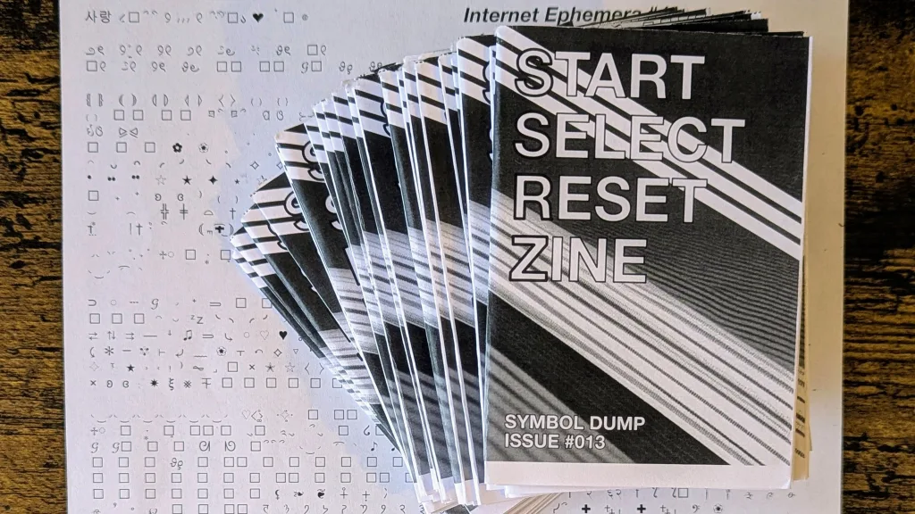 A stack of zines titled START SELECT RESET ZINE Issue #013 Symbol Dump fanned out on a sheet of printed unicode symbols.