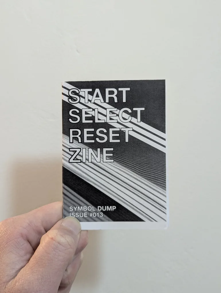 Start Select Reset Zine Issue #013 - a hand holding the small black and white booklet against a plain white wall.