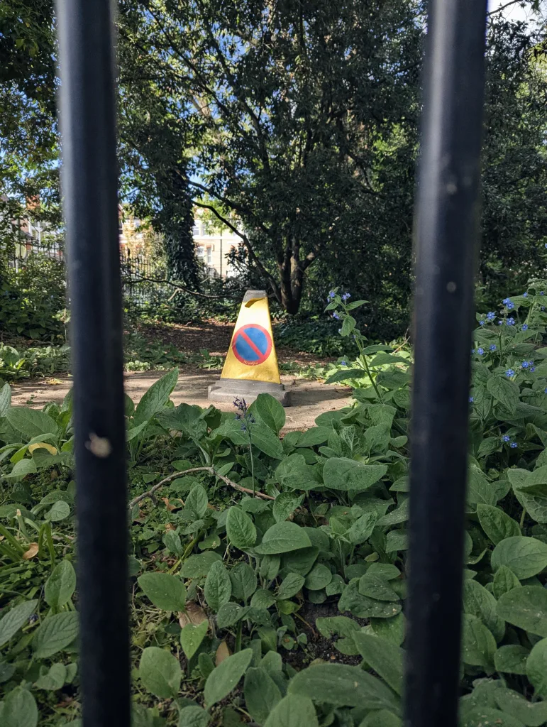 A yellow no-waiting traffic cone on a garden path, framed by vertical black railings and surrounded by lush green foliage.