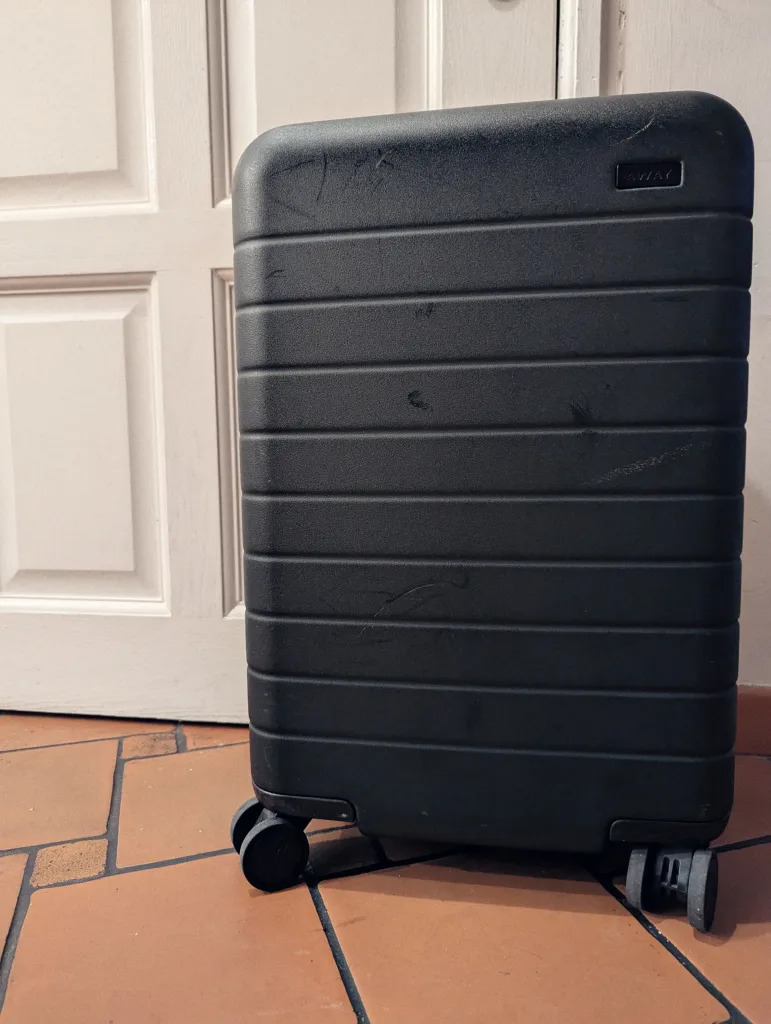 A scuffed black Away hard-shell suitcase with horizontal ridges on a terracotta tiled floor against a white paneled door.