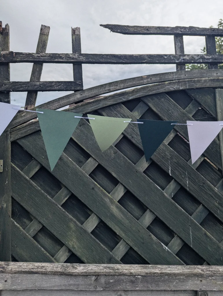 Green and lavender paper bunting strung across a weathered dark wood lattice fence under a cloudy sky.