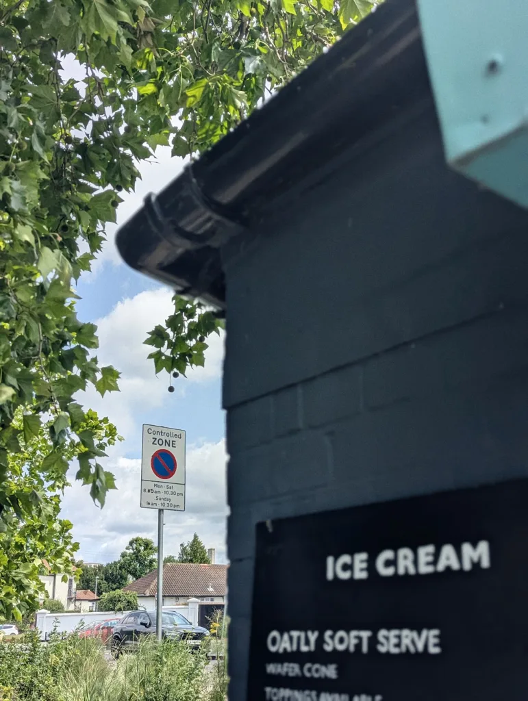 An Oatly soft serve ice cream menu on a dark wall, with a controlled parking zone sign and leafy green trees in the background.