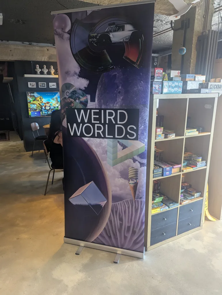 A "WEIRD WORLDS" pull-up banner with surrealist collage art stands in a room beside a wooden shelf filled with board games.