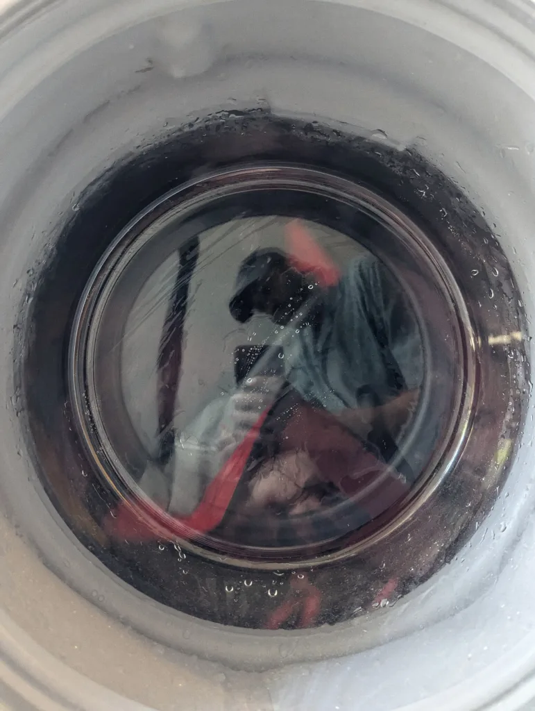 A distorted reflection of a person in a cap taking a photo, visible in liquid at the bottom of a white cup with condensation.