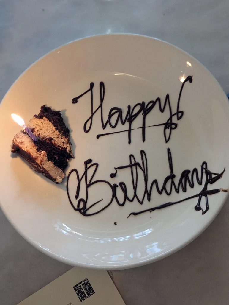 A small slice of chocolate mousse cake with a lit purple candle and Happy Birthday written in chocolate script on a white plate.