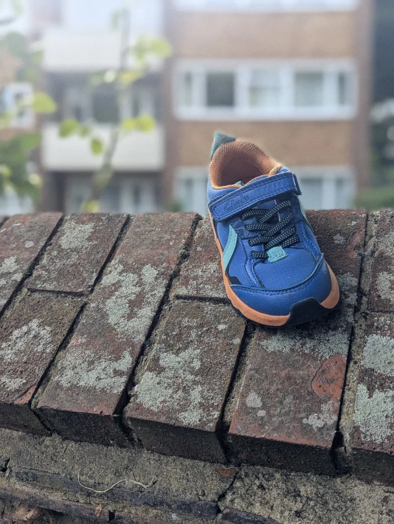 A single blue and orange Clarks toddler sneaker balanced on a lichen-covered brick wall with a blurred building in the background.