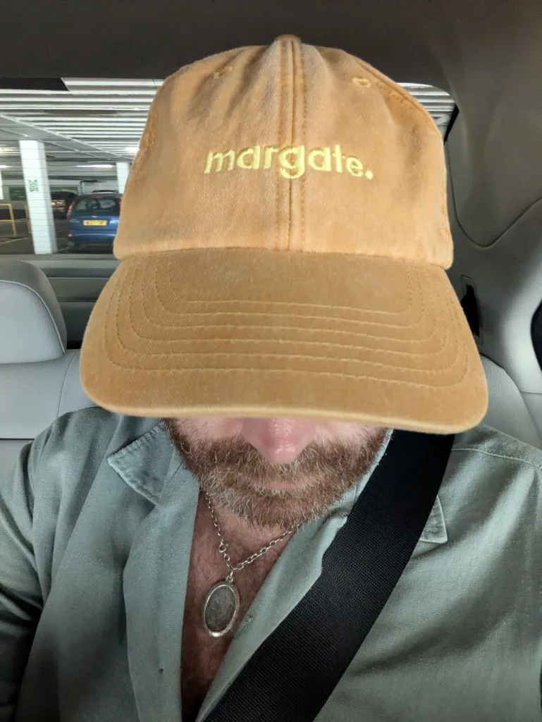 A bearded man in a green shirt wearing a mustard yellow baseball cap with "margate." embroidered on the front, sitting in a car.